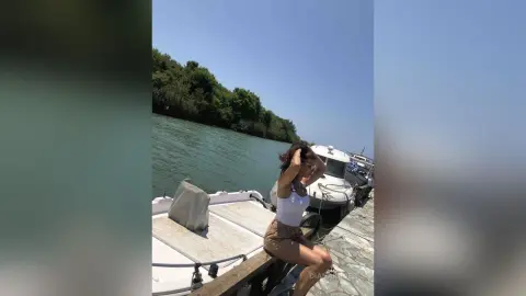 Young woman in a white tank top relaxing on a boat by a scenic river under a clear blue sky.
