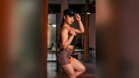 VickyPortman poses in a black bra and brown leather skirt, looking over her shoulder with her hand near her head.