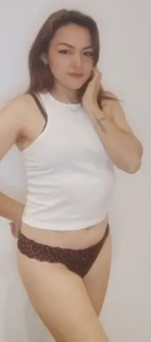 Young woman in a white tank top and brown patterned bikini bottoms posing against a white wall.
