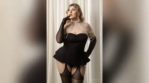 Elegant woman in a black corset and pearl necklace posing glamorously against white curtains.