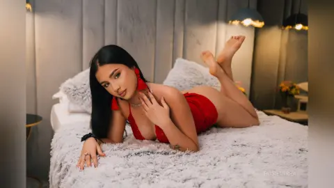 Young woman in a red bodysuit relaxing on a white shag bed in a cozy, dimly lit bedroom.