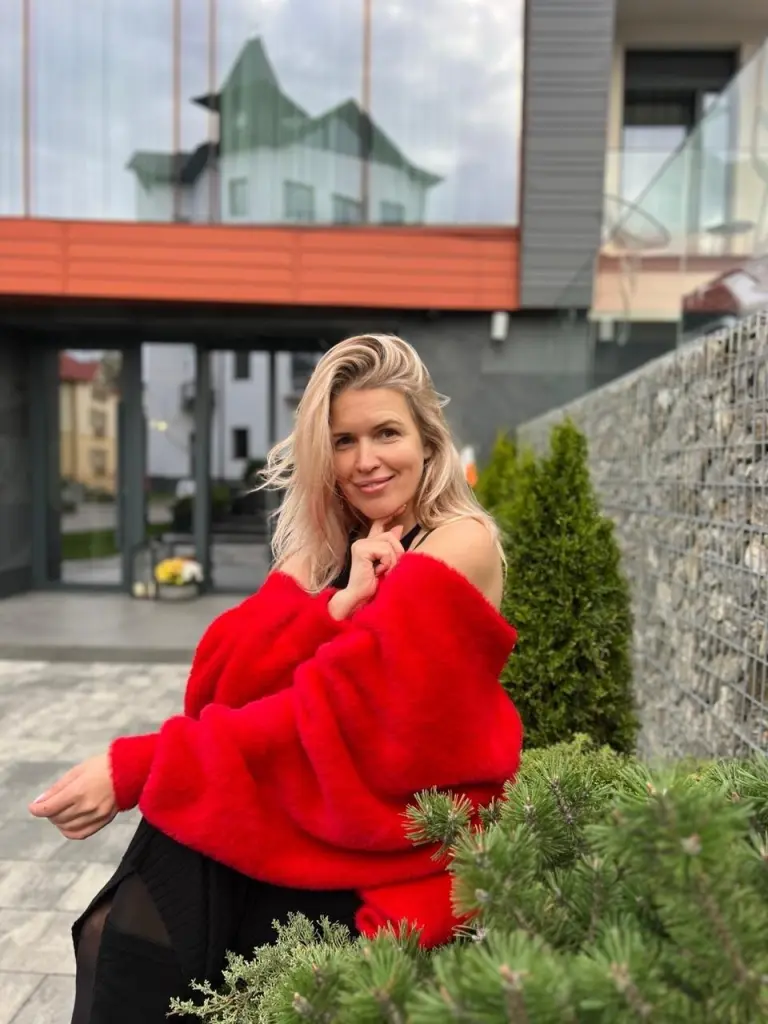 NikaZen is a smiling blonde woman wearing a yellow tank top and denim jacket, posing outdoors next to red flowers.