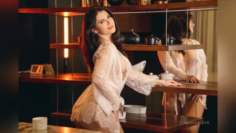 Elegant woman in a white lace sheer robe posing in a warm, moody kitchen setting.
