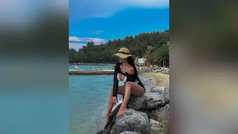 NicoleBella sits on large rocks by the ocean wearing a black mesh bikini and a wide-brimmed straw hat.