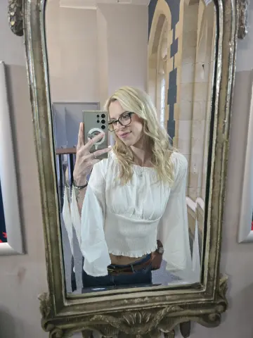 Blonde woman in a white bohemian blouse taking a mirror selfie in an elegant vintage room.