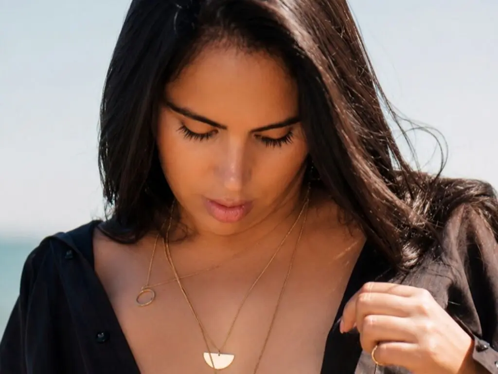 Serene woman with long dark hair wearing a black linen shirt and gold necklaces at a sunny beach.