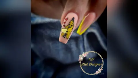 Hand with creative nail art featuring a cute yellow banana and red heart design on a nude base.