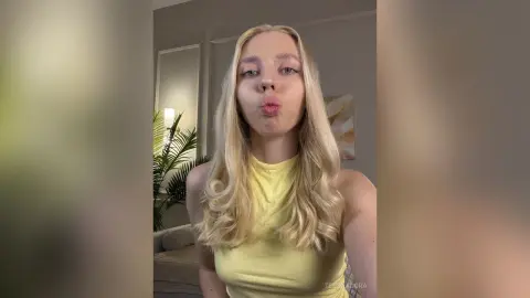 Blonde woman in a yellow sleeveless top making a playful kissy face in a bright indoor setting.