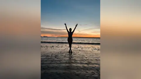 Silhouette of a woman with arms raised on a beach at sunset, enjoying a peaceful ocean view.