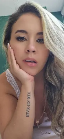 Close-up portrait of a blonde woman in white lace loungewear with a Roman numeral tattoo on her forearm.