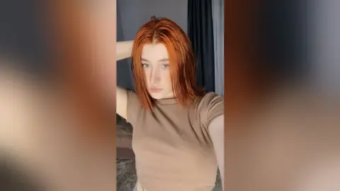 KeithaHall poses with her red hair and one hand behind her head, wearing a tight brown t-shirt.