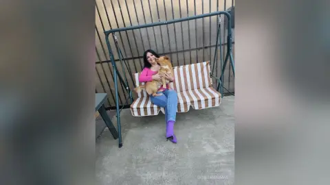 OpheliaDumas smiles while sitting on a striped swing, holding a light brown dog in her lap while wearing a pink top and blue jeans.