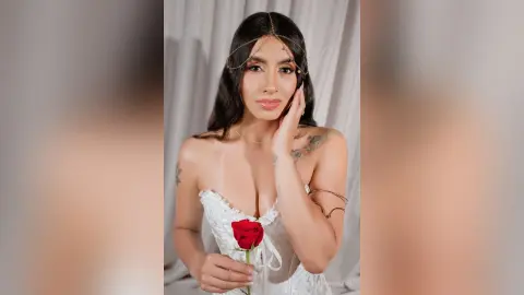 Elegant woman in a white lace corset holding a red rose against a soft white curtain background.