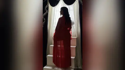 Elegant woman in a flowing red gown standing by a window in a moody, cinematic indoor setting.