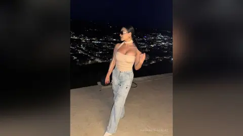 Stylish woman in a beige tube top and light wash jeans posing against a glowing city skyline at night.
