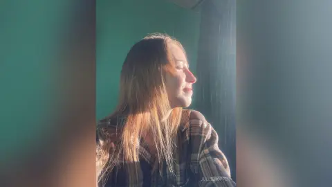 Young woman with long hair wearing a plaid shirt sitting in warm sunlight by a window.