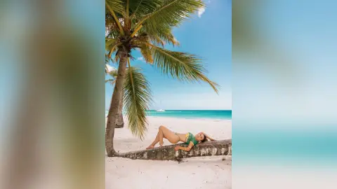 Relaxed woman in a green bikini lounging under a palm tree on a sunny tropical white sand beach.