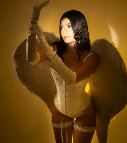 Woman in white corset and long gloves wearing angel wings against a warm golden background.
