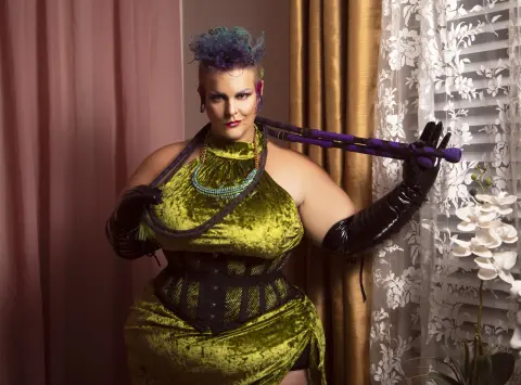 Woman in lime green velvet dress and black corset posing with purple accessories in a warm indoor setting.