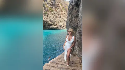 Woman in a white crochet dress standing on stone steps by a turquoise Mediterranean sea and rocky cliffs.