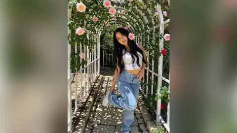 Young woman in white crop top and blue jeans posing in a sunny rose garden walkway.
