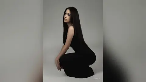 Elegant woman in a sleek black dress posing against a minimalist grey background with a sophisticated mood.
