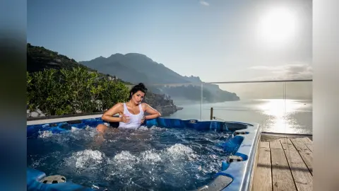 MissyJolie relaxes in a bubbling hot tub overlooking a coastal mountain view, wearing a white tank top.