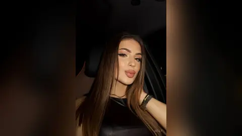 Glamorous woman with long brown hair and winged eyeliner posing in a dark car at night.