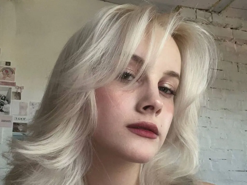 Close-up portrait of a blonde woman with wavy hair and natural makeup in a bright, indoor setting.