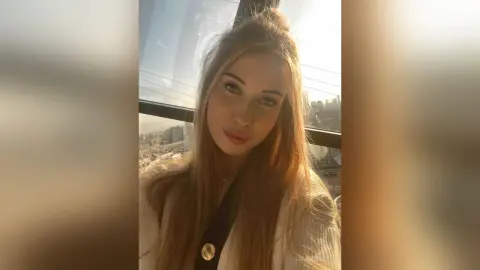 Young woman with long blonde hair wearing a white sweater in a sunlit high-rise apartment overlooking a city.