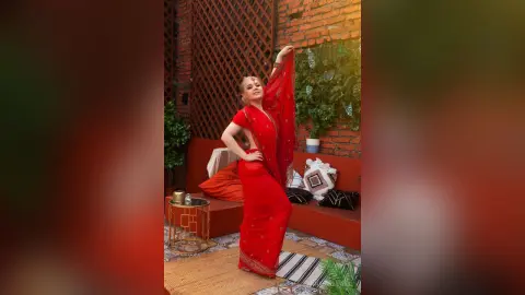 Elegant woman in a vibrant red traditional saree posing gracefully in a cozy, sunlit indoor garden setting.