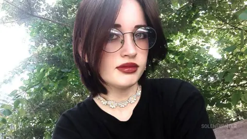 SoleneWinter wearing round glasses and a black t-shirt and a white flower choker, posing against a background of green leaves.