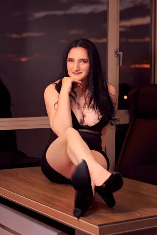 Smiling woman in a black dress sitting on a desk in a modern office at sunset.