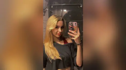 Blonde woman in a black crop top taking a mirror selfie in a modern bathroom with warm lighting.