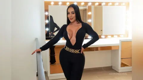 Smiling brunette woman in a sleek black bodysuit posing in a bright vanity dressing room.