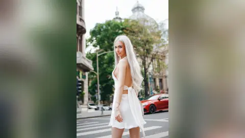 Elegant woman in a white backless dress posing on a city street with classic architecture background.