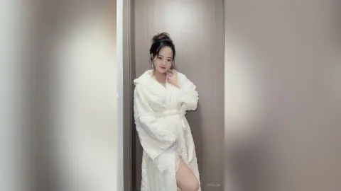 EnyaLin poses in a white fluffy robe against a light wood door, running her hand near her face.