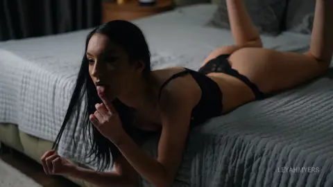 Young woman with long dark hair in black lace lingerie lying on a bed in a moody, dimly lit bedroom.
