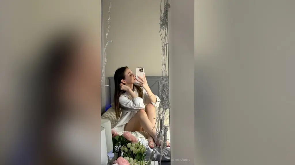 AlthenaBute — Young woman in a white long-sleeve shirt taking a mirror selfie in a cozy, dimly lit bedroom.