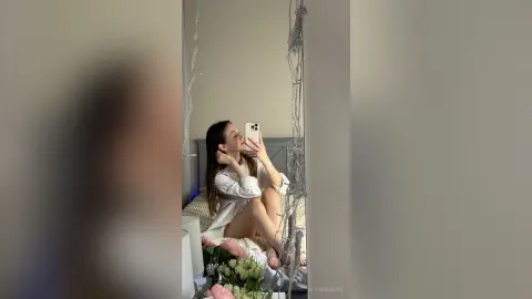 Young woman in a white long-sleeve shirt taking a mirror selfie in a cozy, dimly lit bedroom.