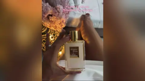 AnaMathers holds a bottle of Kilian perfume near her bare legs and feet, surrounded by soft pink flowers.
