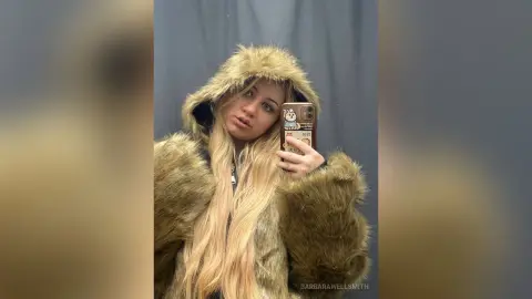 Blonde woman in a cozy brown faux fur hooded coat taking a selfie against a dark background.
