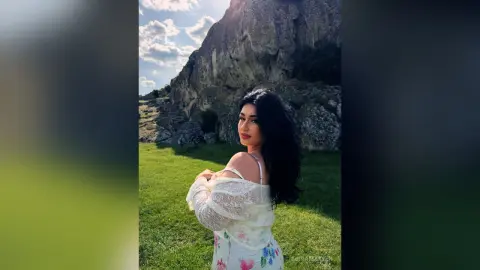 Woman in white floral lace dress posing in a sunny mountain meadow with rocky cliffs.