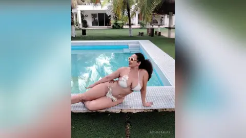 Curvy woman in a white bikini and sunglasses relaxing by a sunny outdoor swimming pool.