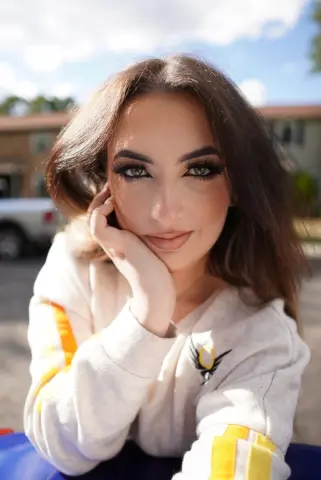 Smiling woman in a white sweatshirt relaxing outdoors under a bright sunny sky.