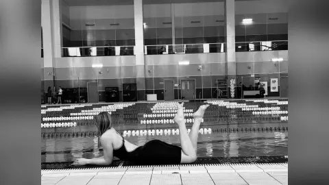 Woman in black swimsuit lying on poolside edge of an indoor swimming pool in a moody black and white photo.
