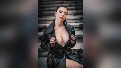 Glamorous woman in a black leather trench coat posing on stone stairs with a bold, moody aesthetic.
