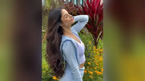 TefyRusso poses outdoors with her long, wavy dark hair flowing down her back, wearing a light blue zip-up hoodie and white crop top.