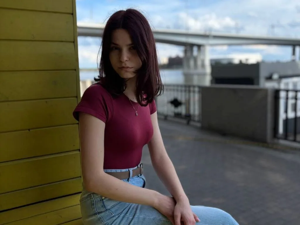 KrystalBeadling wears a tight maroon t-shirt and blue jeans while leaning against a yellow wall outdoors.