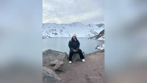 Woman in black winter jacket and pink beanie sitting on rocks overlooking a snowy mountain lake landscape.